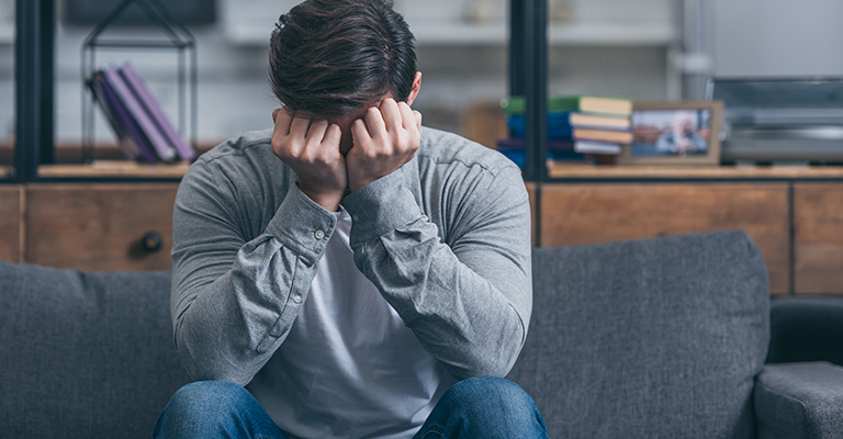 man sitting on couch, crying and and covering face with hands at home