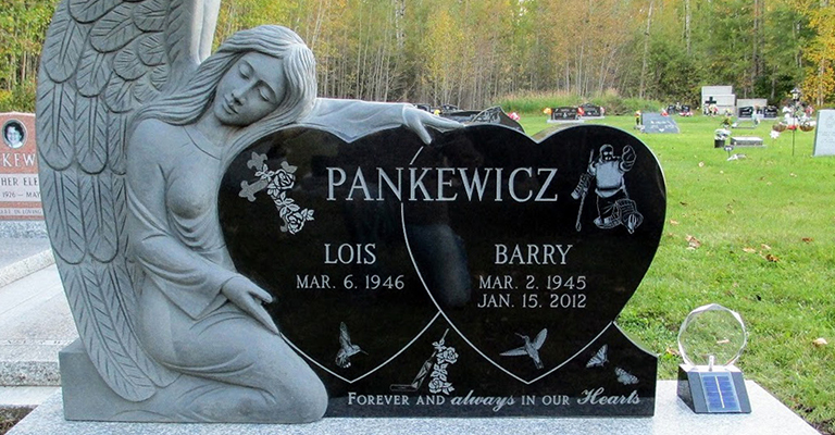Gravestone marker at the cemetery with a large angel beside the marker. The headstone is two hearts with each loved one in their own heart. There is a goalie, a cross, doves, a goalie stick and the etching that says Forever and always in our hearts.