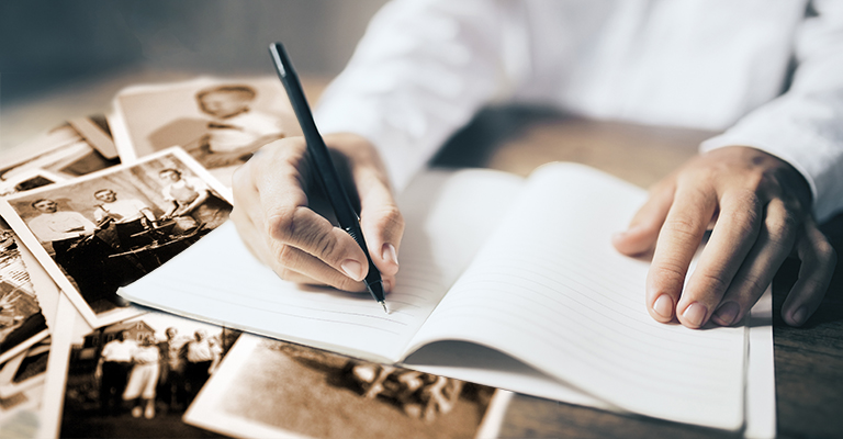 Man writing in blank diary and paper coffee cup on wooden table with old photos on table.