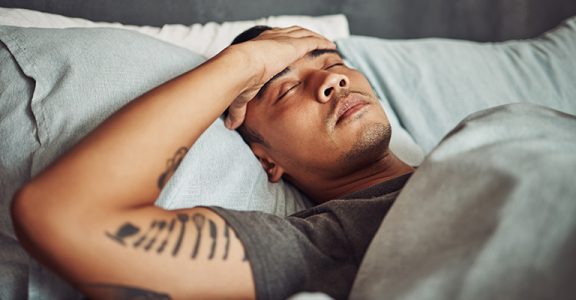 Tired, rest and sick man sleeping in the bedroom while in recovery or healing in his apartment. Burnout, illness and male person with a headache or fever taking a nap in bed at his modern home